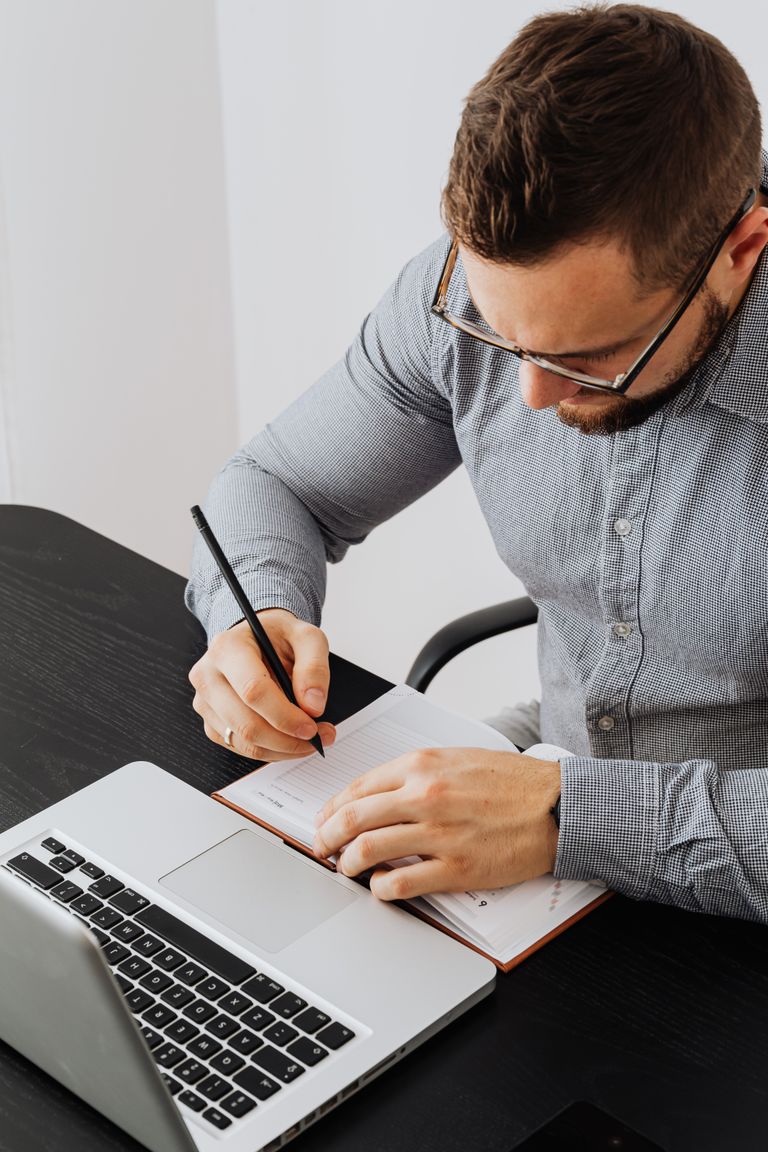 Businessman writing notes on a notebook in a modern office setting with laptop on desk.